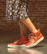 A woman in a floral dress is standing on a brick wall in a pair of orange high top sneakers called Rossela by Bed Stu.