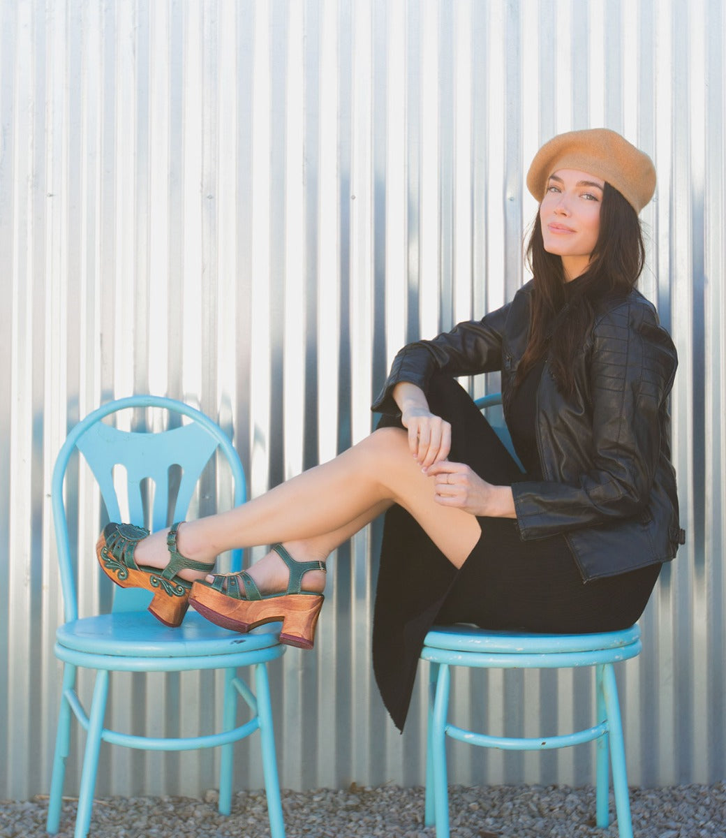A woman wearing tan hat, black jacket and skirt with dark teal Sabine sandals while sitting on blue chairs.