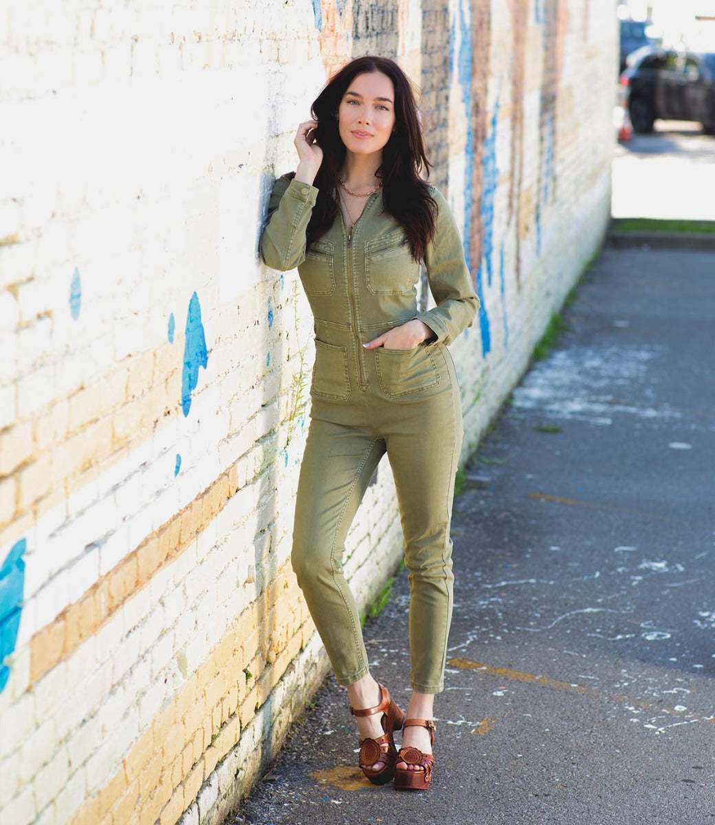 A woman in a green jumpsuit and tan wooden sandals while leaning against a wall.