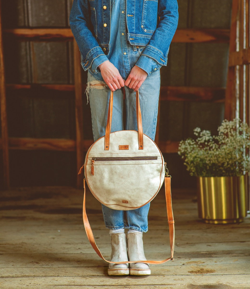A woman wearing a denim jacket and jeans holding a Bed Stu Saray white leather purse.