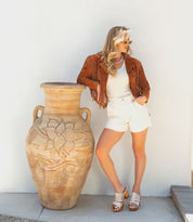 A woman in a brown fringe jacket and white shorts leans against a large decorative clay vase, standing on a stone floor by a white wall, her feet clad in Bed|Stü’s Shantel clogs with woven uppers and cushioned leather insoles.