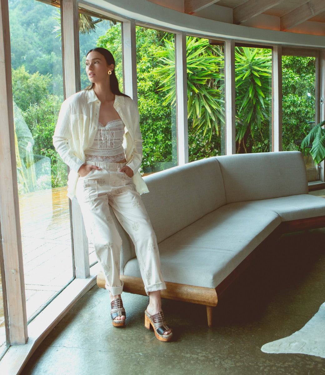 A woman stands indoors by a curved sofa, hands in her pockets, wearing a light-colored shirt, corset top, and pants. On her feet are the cheerful Shantel clogs from Bed|Stü, featuring a woven upper and cushioned leather insole. Large windows reveal lush greenery outside.