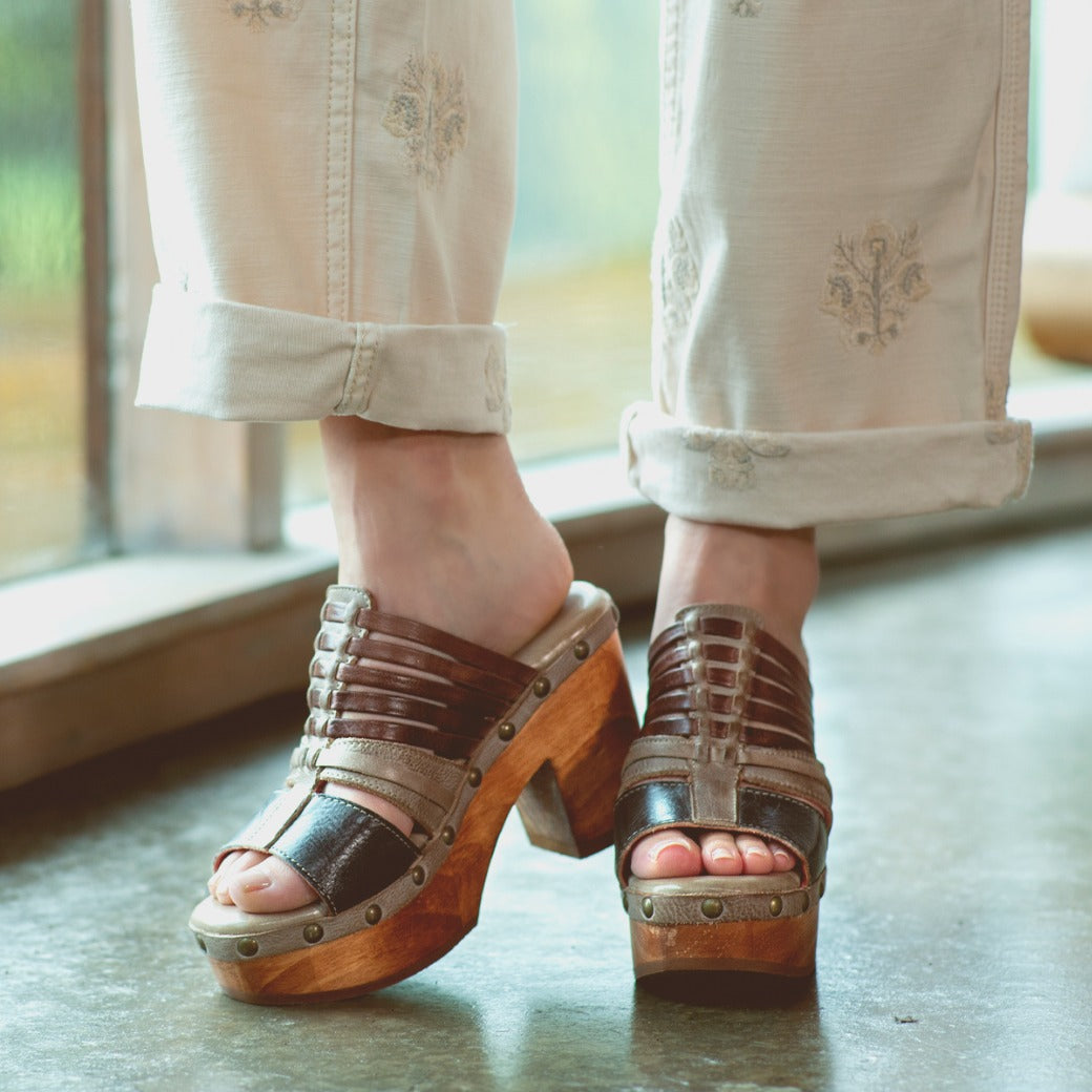 A person wearing cream embroidered pants and the Bed|Stü Shantel brown cheery clog sandals, featuring a cushioned leather insole, stands indoors on a polished floor near a window.