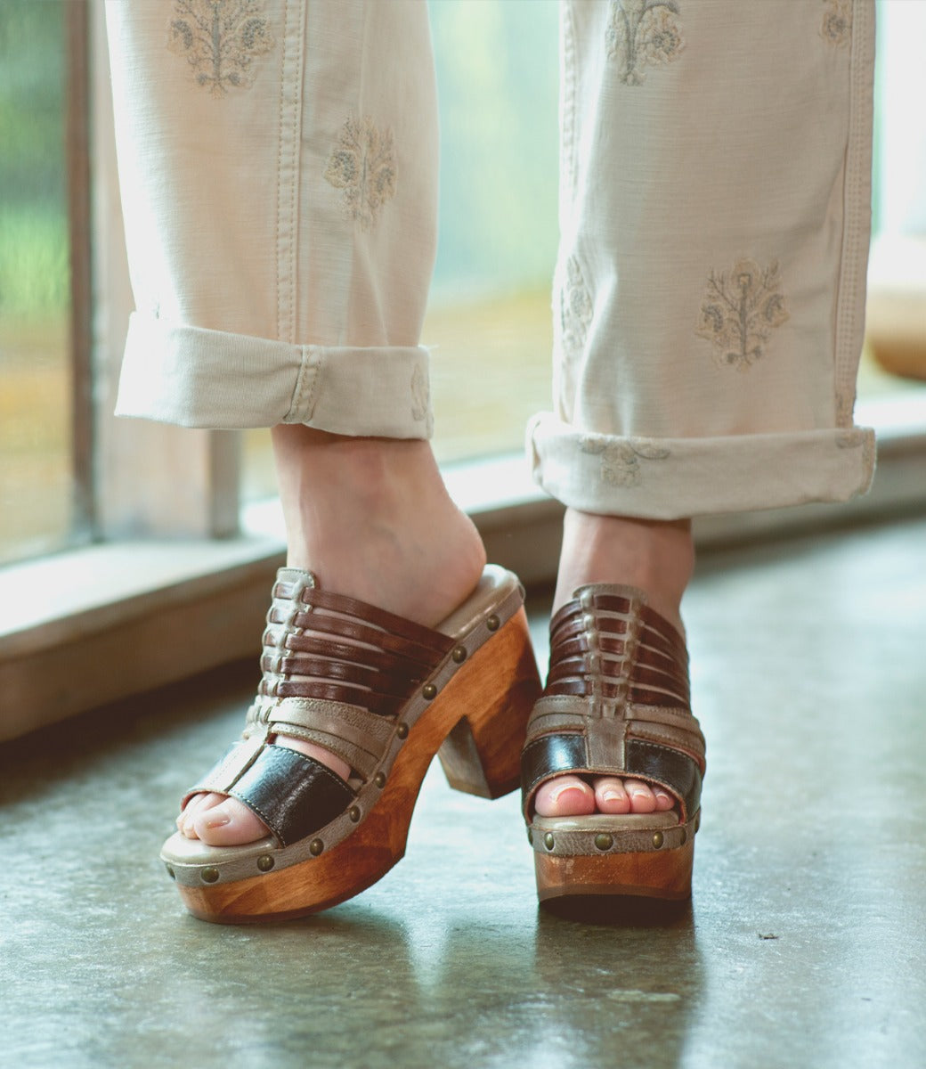 A person wearing cream embroidered pants and the Bed|Stü Shantel brown cheery clog sandals, featuring a cushioned leather insole, stands indoors on a polished floor near a window.
