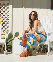 A woman in a white top and colorful patchwork pants sits on a white chair outdoors, wearing sunglasses and holding a small brown clutch. With cacti and a white fence behind her, she completes her look with Bed|Stü’s Shantel clogs, featuring a woven upper and cushioned leather insole for all-day comfort.