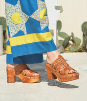 A person wearing the Bed|Stü Shantel brown platform sandals, which feature a woven upper and cushioned leather insole, paired with a blue, yellow, and floral-patterned dress stands on a concrete surface with cactus plants in the background.
