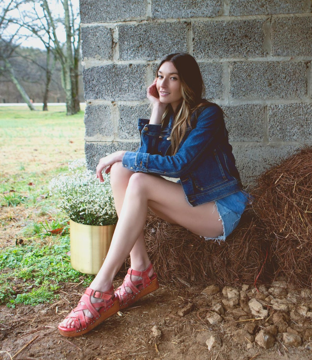 A woman wearing a Bed Stu Shirley denim jacket and shorts sitting on hay.
