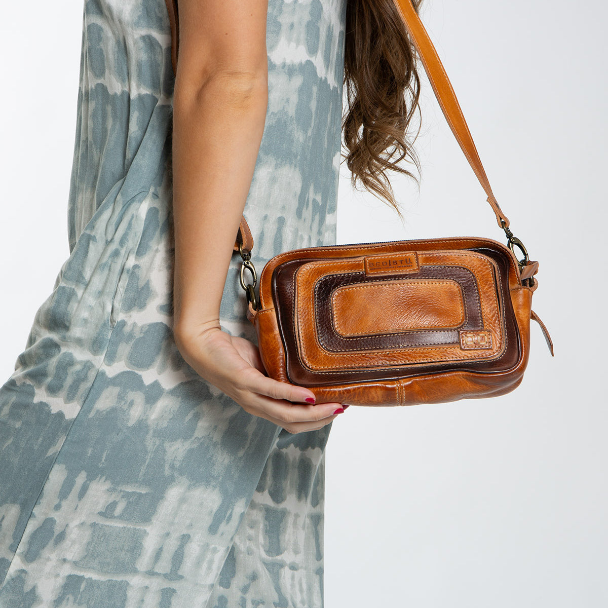 A person wearing a tie-dye dress holds the Shy crossbody bag from Bed Stu, featuring contrasting leather details.