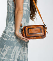 A person wearing a tie-dye dress holds the Shy crossbody bag from Bed Stu, featuring contrasting leather details.
