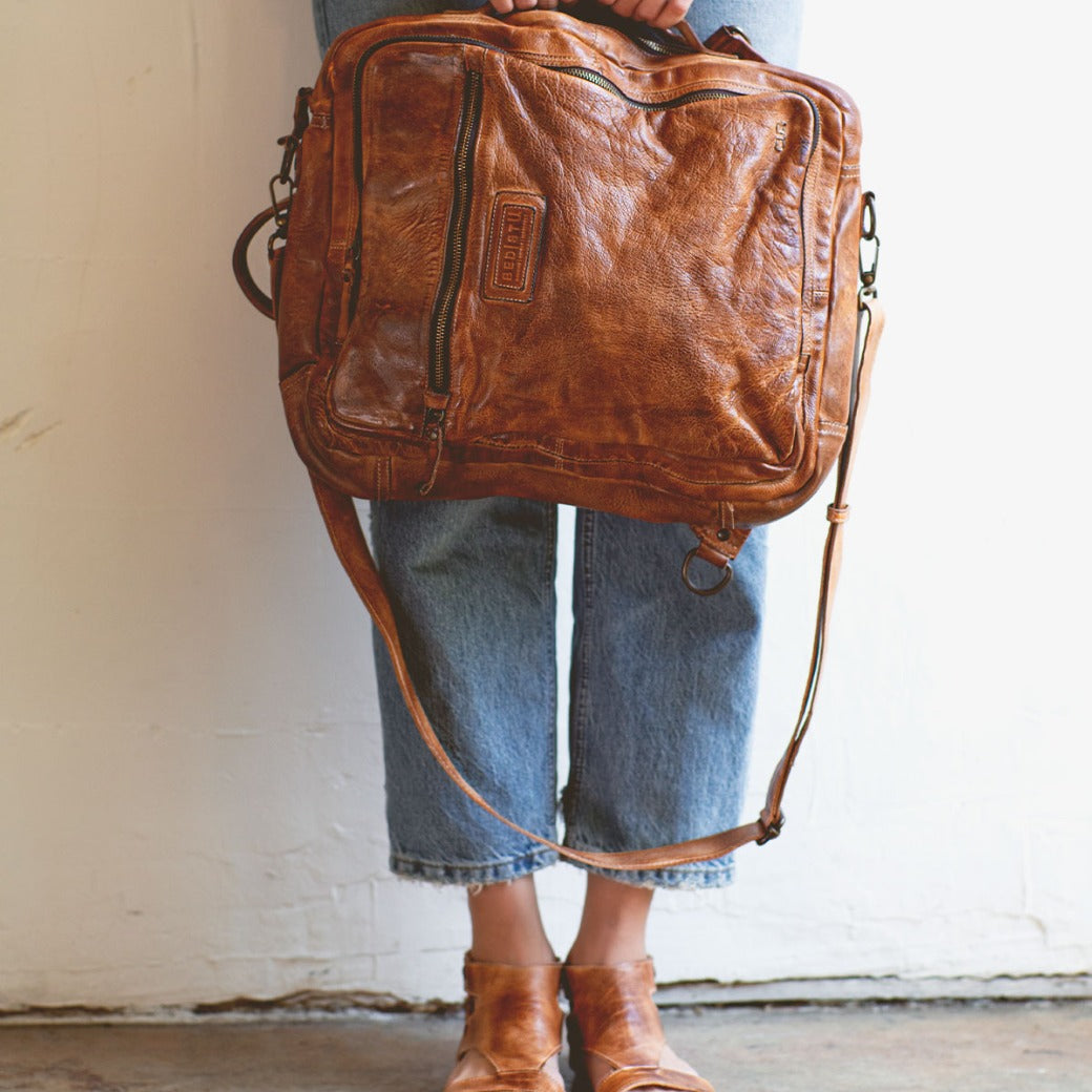 A woman wearing jeans and sandals holding a Bed Stu Socrates brown leather bag.