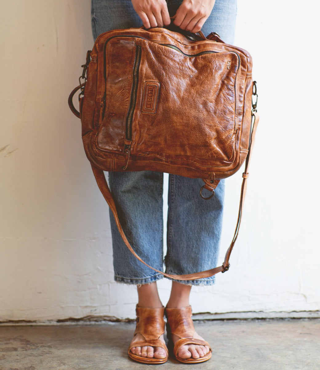 A woman wearing jeans and sandals holding a Bed Stu Socrates brown leather bag.