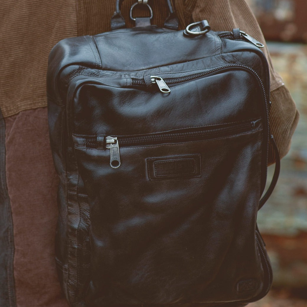 A man is holding a black leather Bed Stu Socrates backpack.
