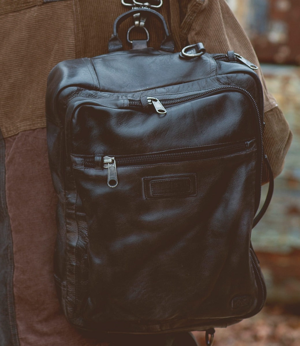 A man is holding a black leather Bed Stu Socrates backpack.