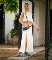 A woman stands outdoors wearing the cream-colored Soto sleeveless top and skirt set from Bed Stu, paired with teal cushioned footbed sandals and a teal crossbody bag—an ideal look for everyday exploring. She faces forward with one hand slightly raised.