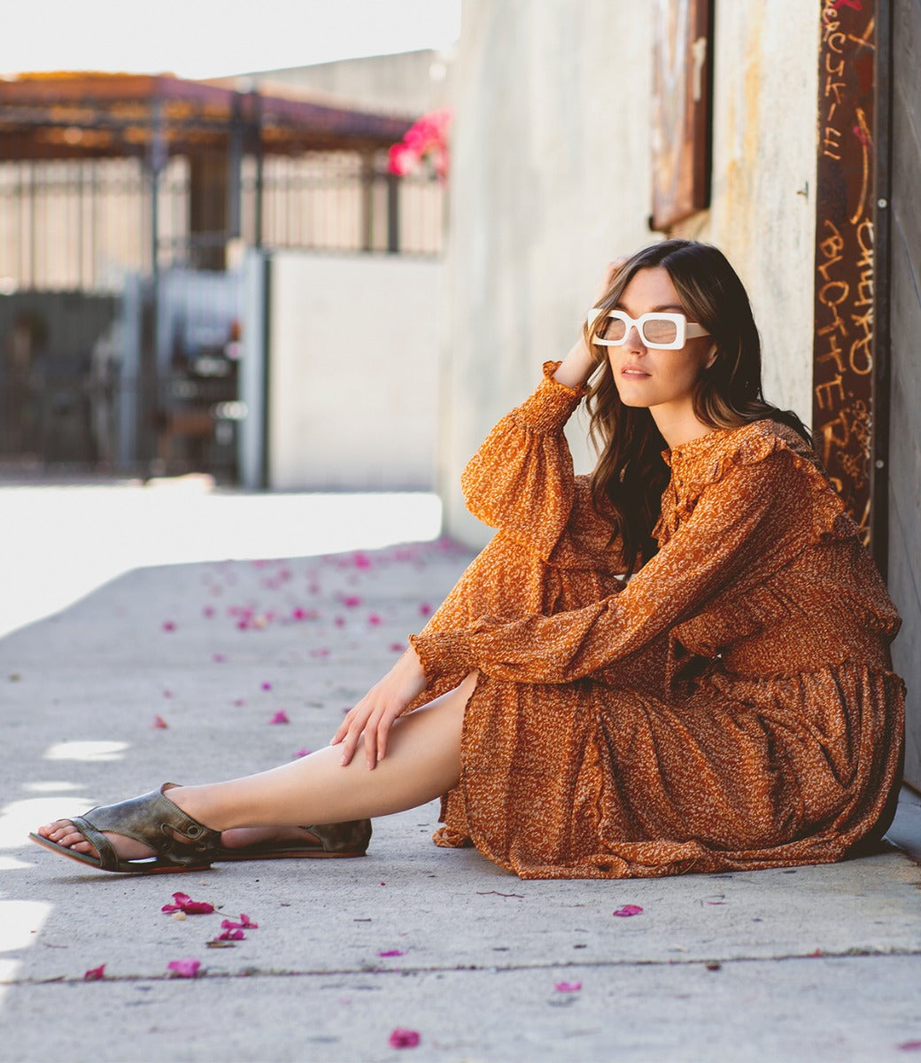 A woman sitting on the sidewalk wearing Bed Stu sandals, sunglasses and a tan polka dot dress.