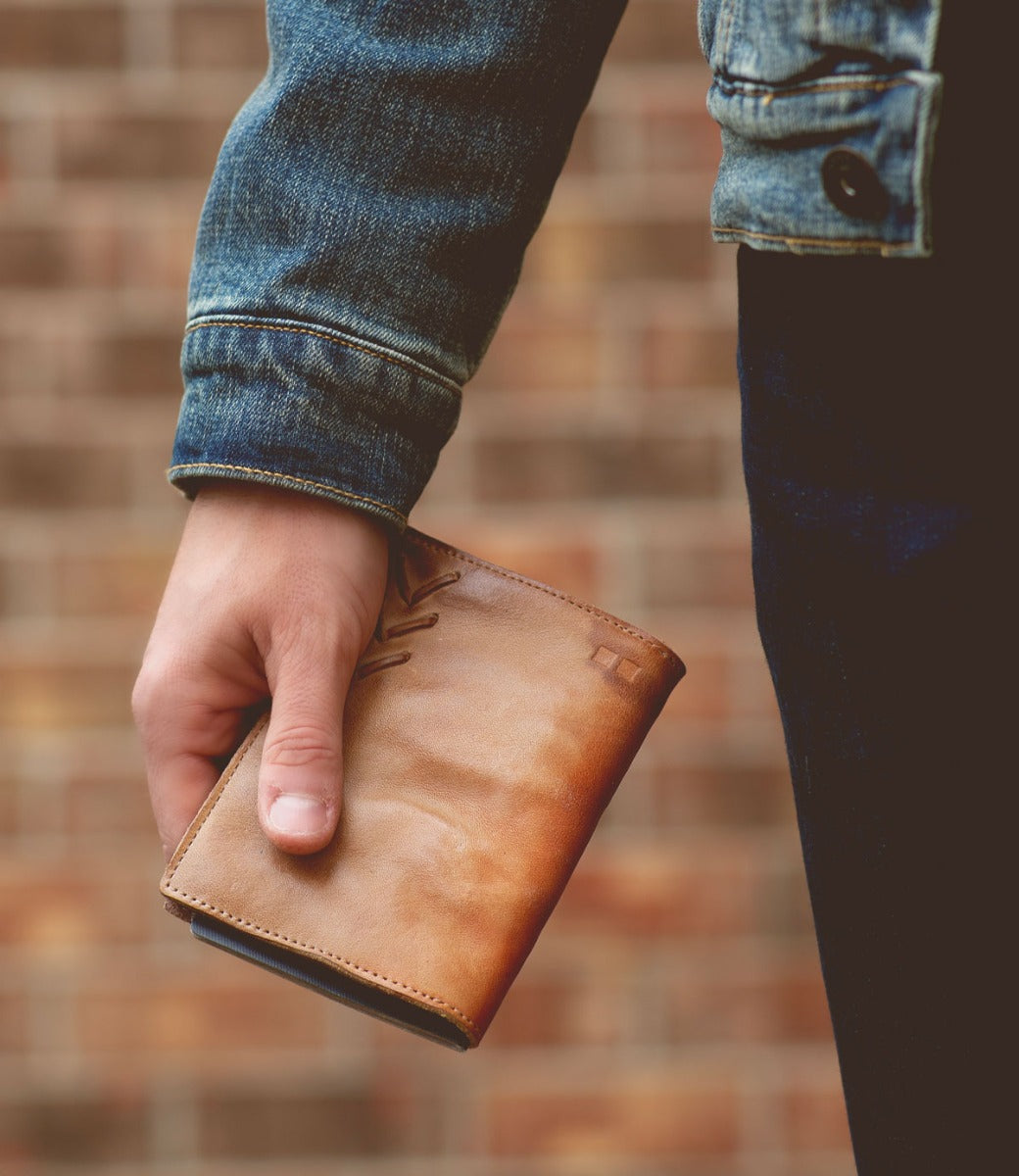 A man holding a Bed Stu Stardust brown leather wallet in front of a brick wall.