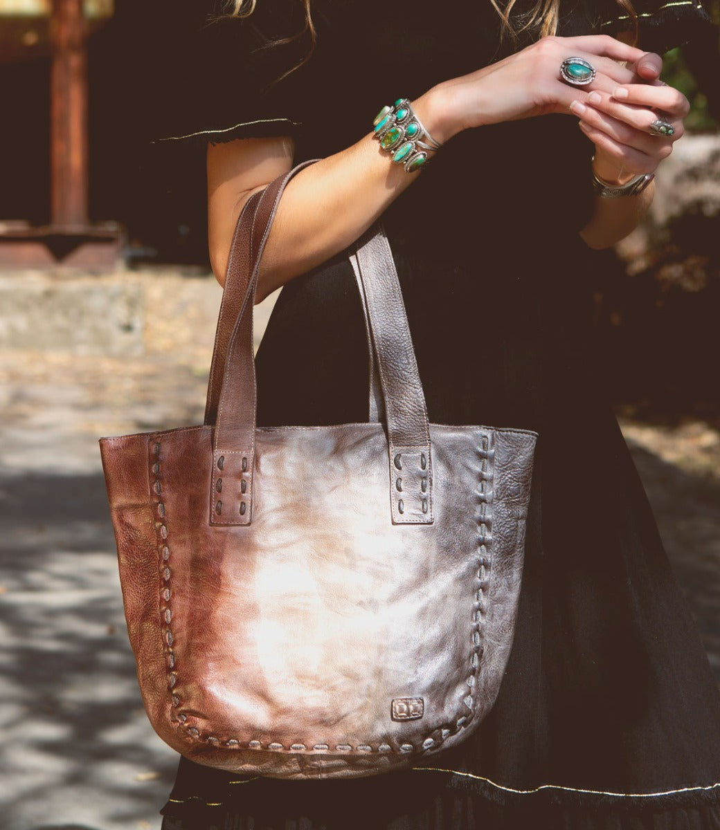 A woman is holding a brown leather Bed Stu Stevie tote bag.