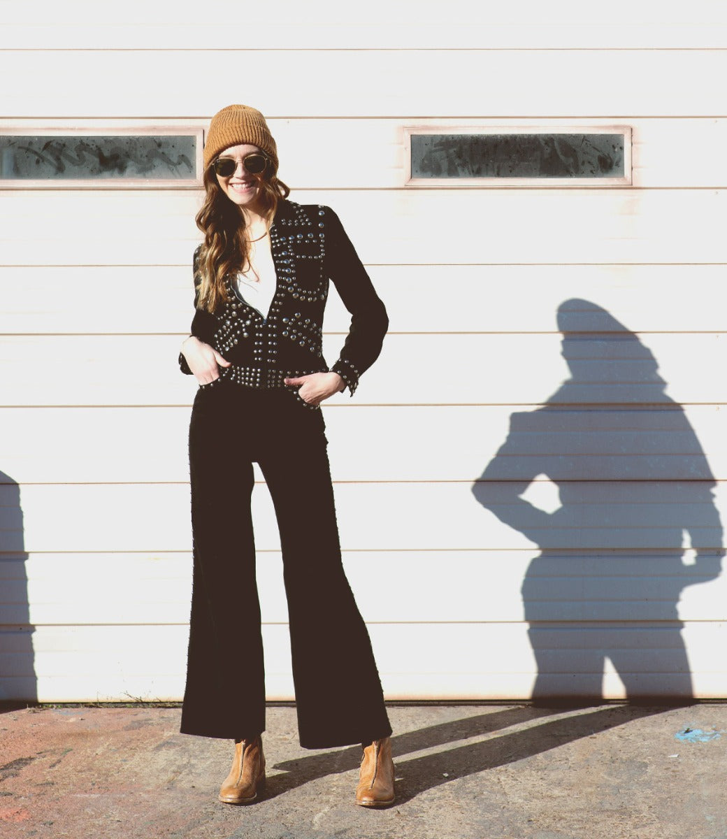 A woman is standing in front of a garage wearing black pants and a beanie from Bed Stu.