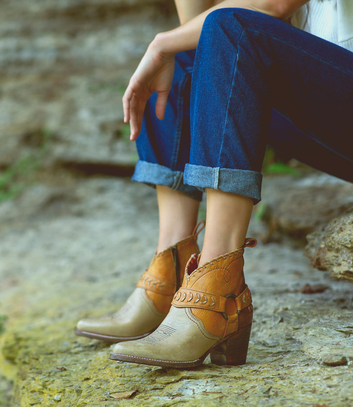 A woman wearing Tania jeans and Bed Stu cowboy boots sitting on a rock.