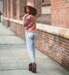 A woman wearing a Bed Stu pink sweater and jeans standing on a brick wall.