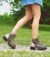 A woman is walking on a dirt road in a pair of Bed Stu Tanzania boots.