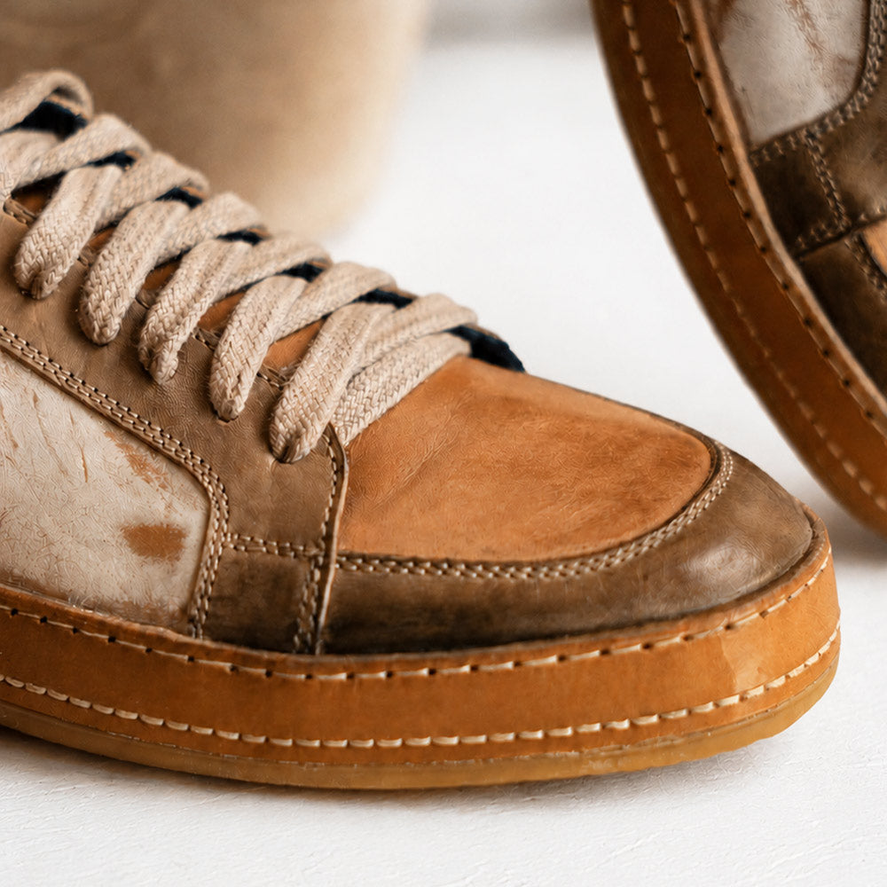 Close-up of a brown and tan lace-up sneaker with cream laces, visible stitching, and a thick gum-colored sole.