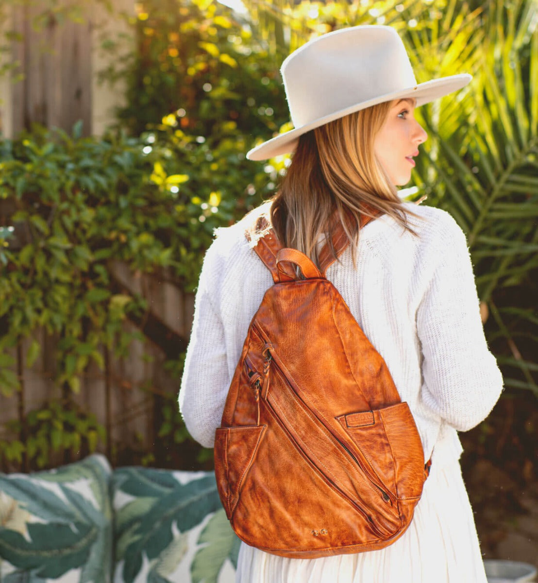 A woman wearing a white hat and a brown leather Tommie backpack by Bed Stu.