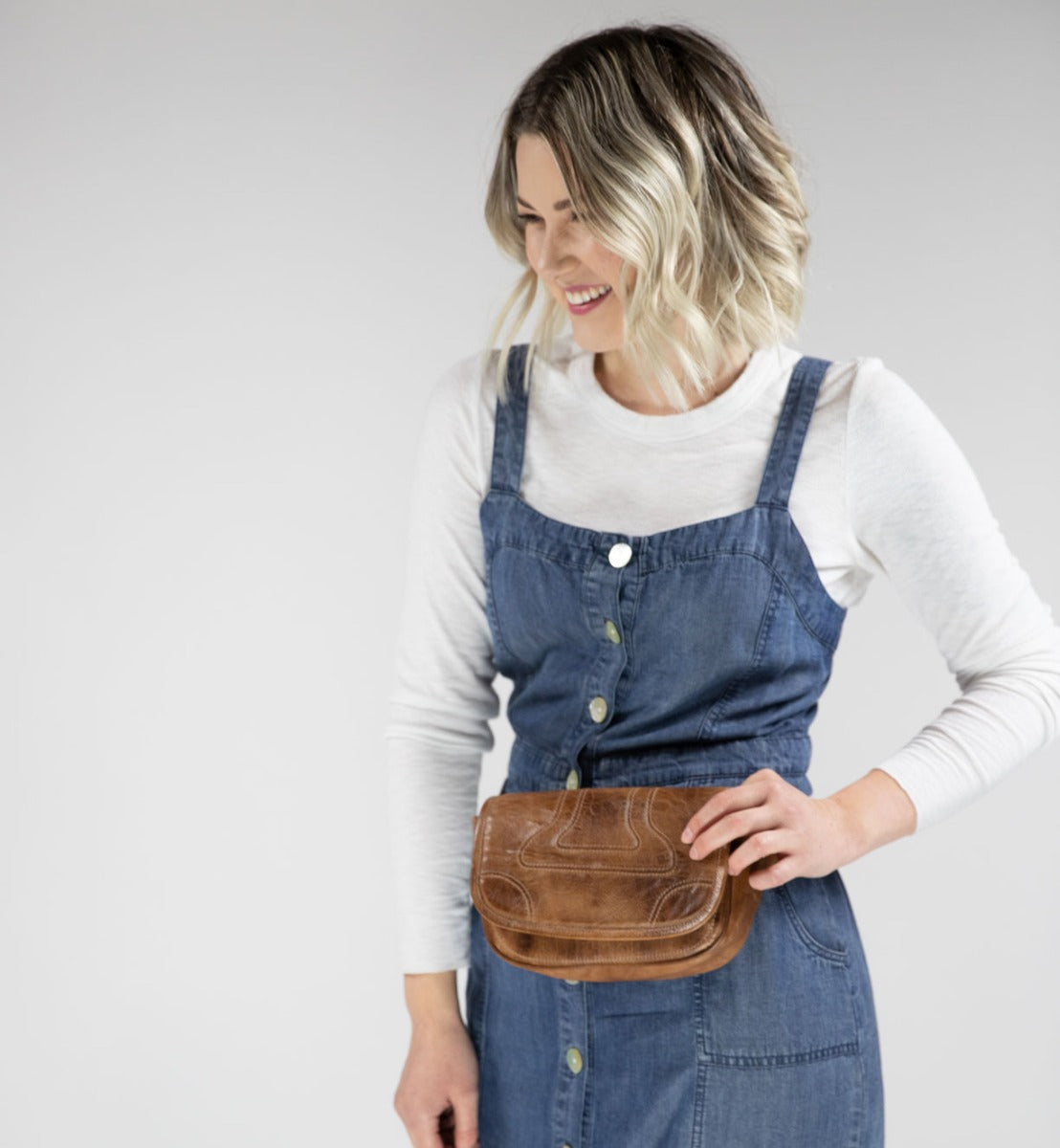 A woman wearing a Travers denim overall dress and Bed Stu belt bag.