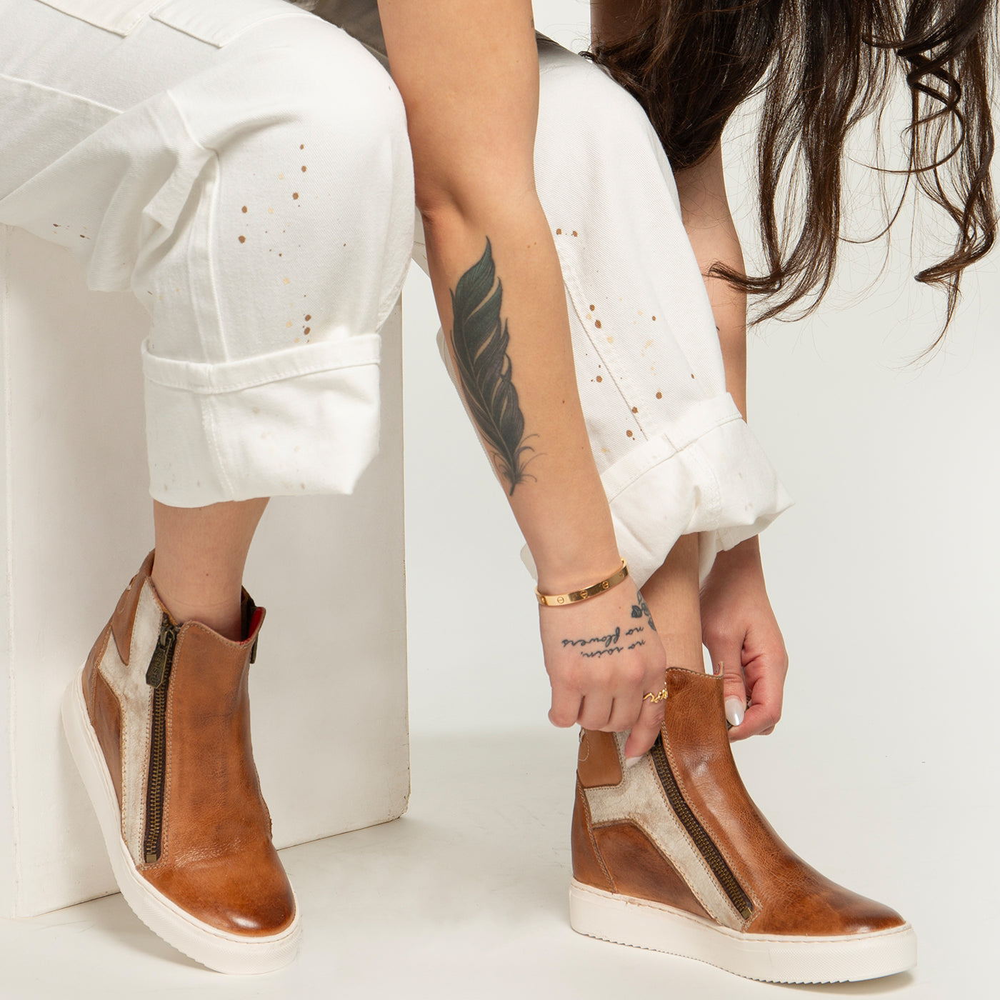 A person with long hair and a leaf tattoo adjusts Bed Stu’s Twist brown leather high-top sneakers, which feature hidden wedge and zipper details, while wearing white pants.