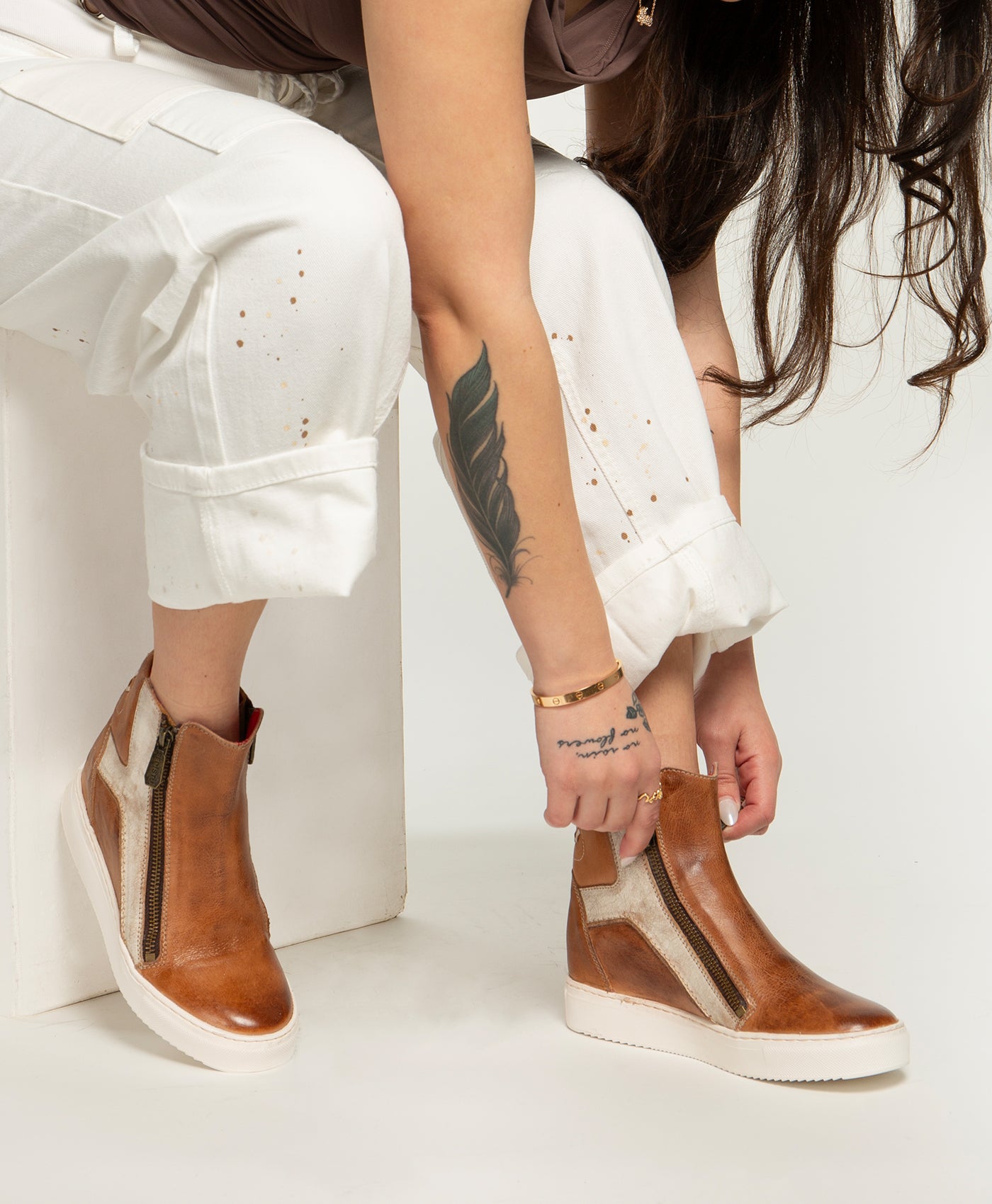 A person with long hair and a leaf tattoo adjusts Bed Stu’s Twist brown leather high-top sneakers, which feature hidden wedge and zipper details, while wearing white pants.