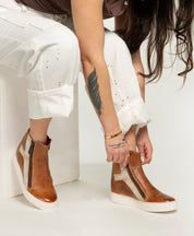 A person with long hair and a leaf tattoo adjusts Bed Stu’s Twist brown leather high-top sneakers, which feature hidden wedge and zipper details, while wearing white pants.