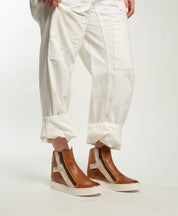 A person wears Bed Stu Twist brown high-top sneakers with side zippers and leather panels, paired with white pants, standing against a plain white background.