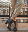 A pair of Isla black leather heeled booties paired with rolled-up beige trousers, against an urban fence backdrop.