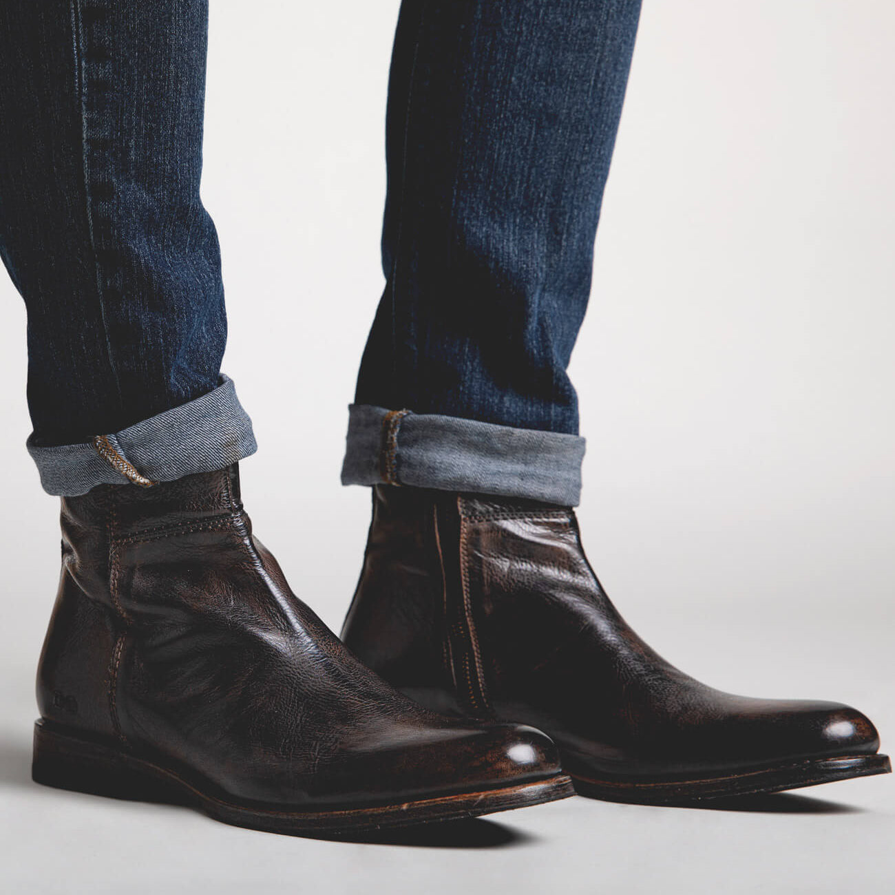 A person wearing Bed Stu Kaldi dark brown leather ankle boots and blue jeans with cuffed hems, standing against a light gray background.