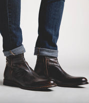 A person wearing Bed Stu Kaldi dark brown leather ankle boots and blue jeans with cuffed hems, standing against a light gray background.