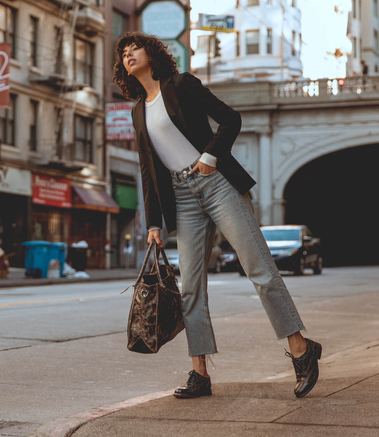 A woman wearing black jacket, white shirt, jeans and black lita shoes while holding a BedStu bag on the street.