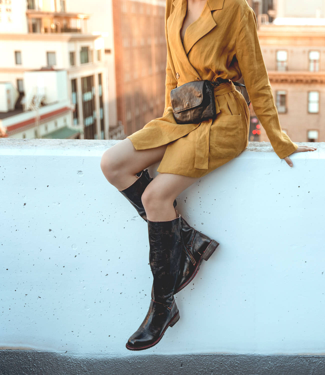 A woman in a yellow coat and black boots sitting on a Manchester ledge Bed Stu.