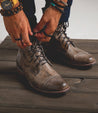 A man is tying his Protégé shoes on a wooden floor.