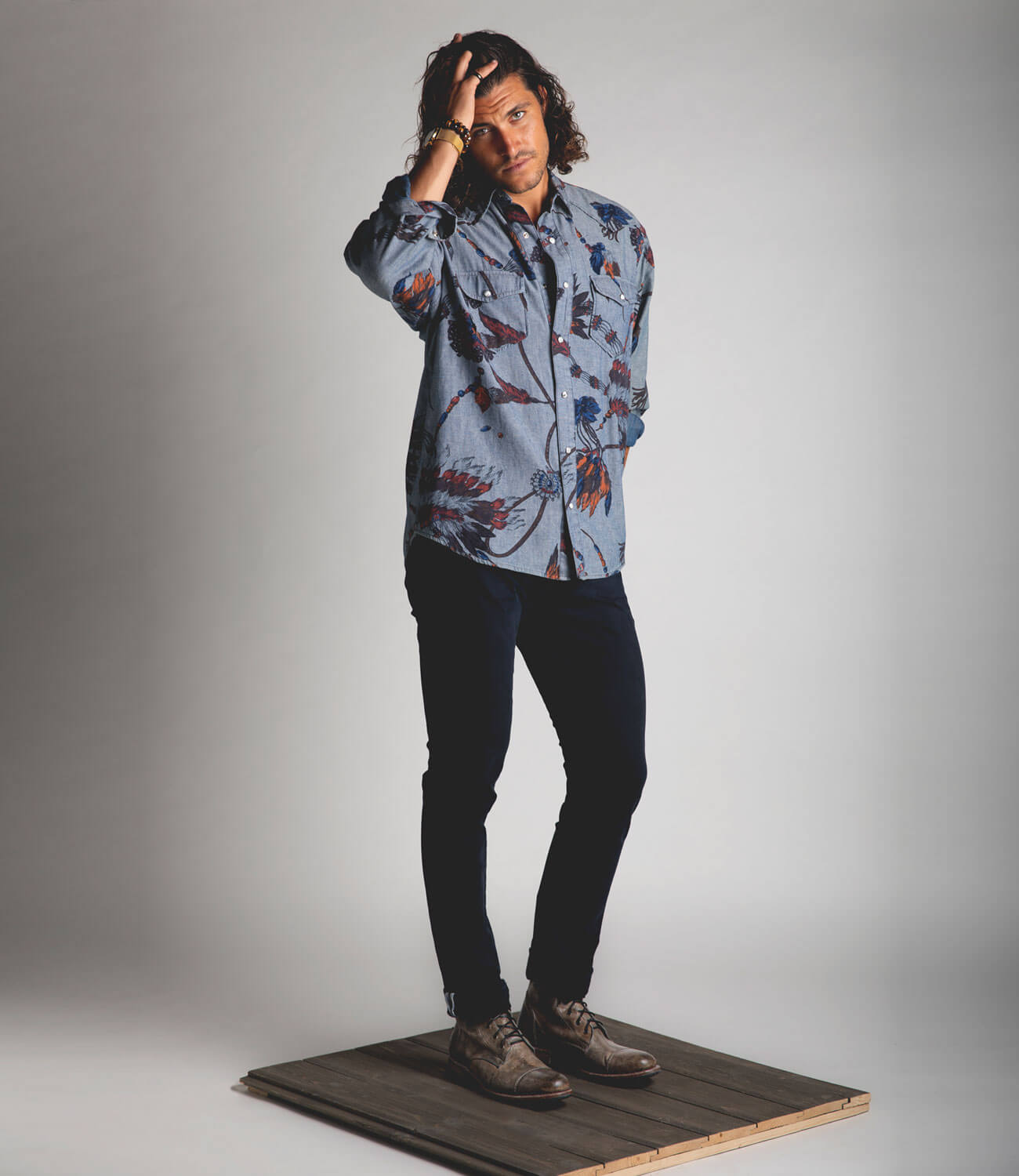 A man in a floral shirt, blue jeans, and Bed Stu Protege boots while standing on a wooden platform.