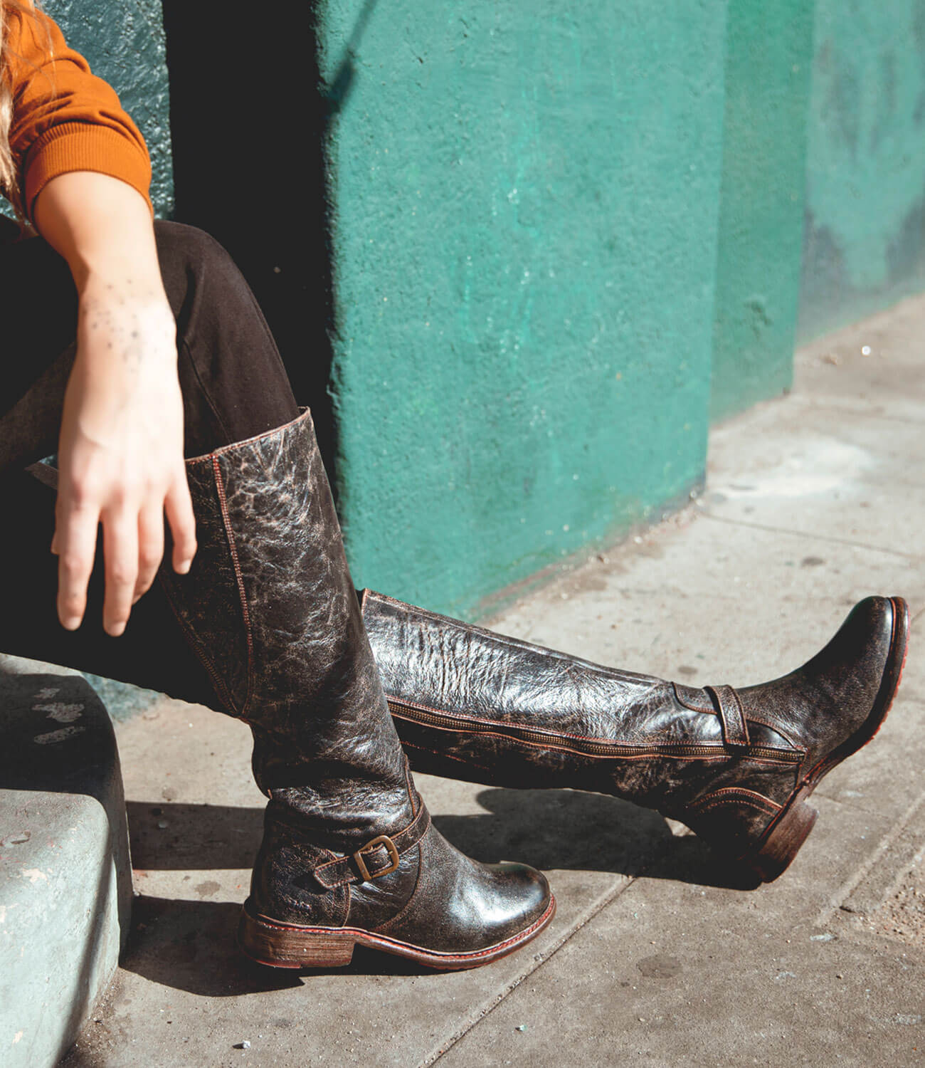 A woman is sitting on a curb in a pair of Bed Stu Glaye boots.