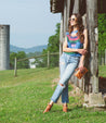 A woman is leaning against a barn in a field, wearing Voleta sandals by Bed Stu.