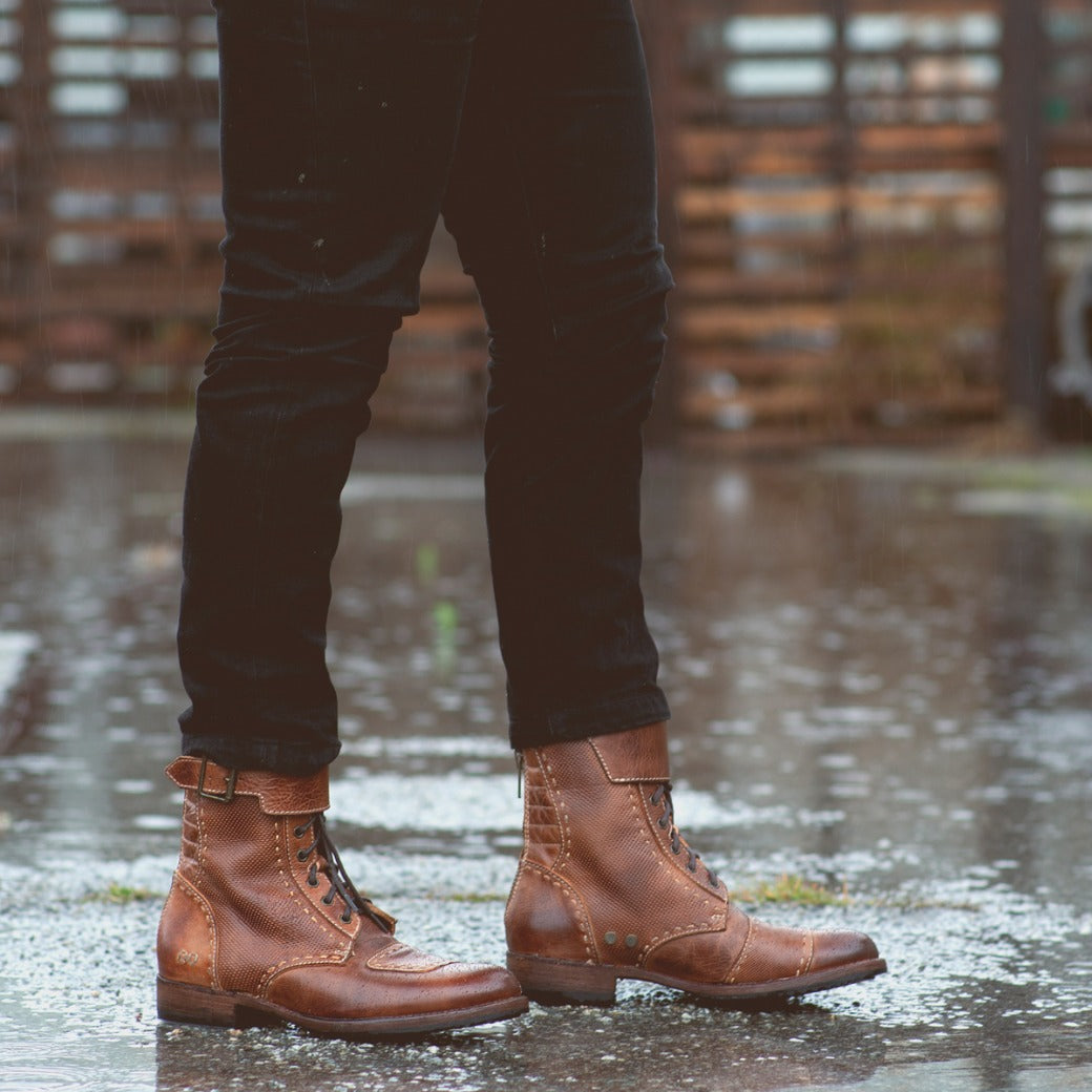 A person wearing Bed Stu's brown leather moto-inspired boots, "Walker," and black jeans standing on a wet surface with puddles, reflecting a cloudy sky.
