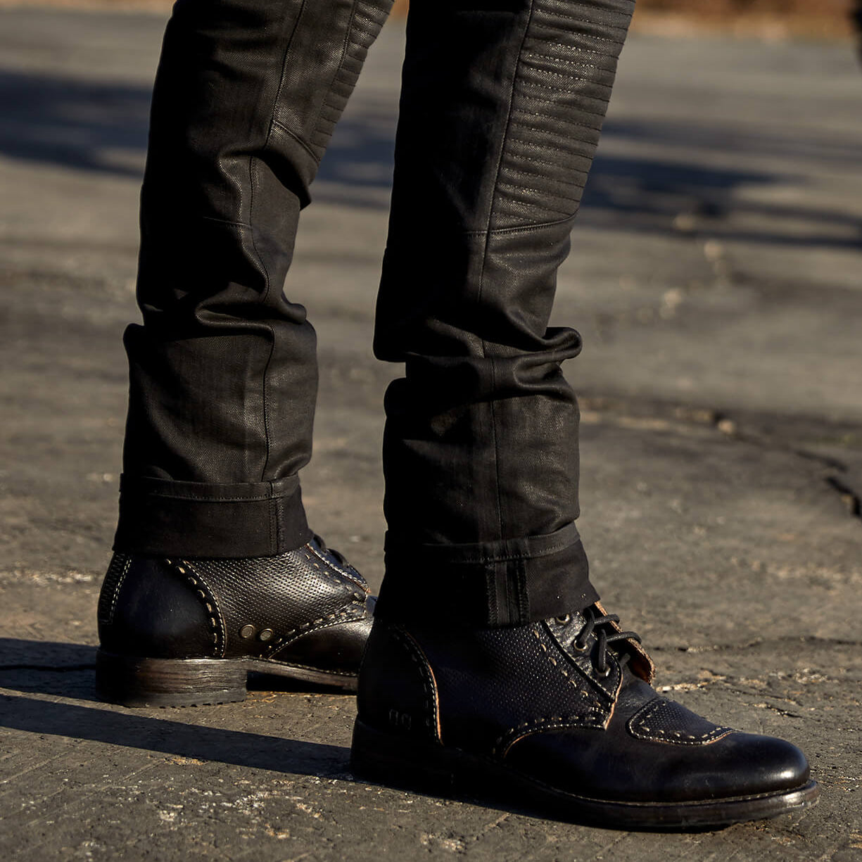 Close-up of a person wearing Bed Stu moto-inspired boots and black jeans standing on a cracked pavement.