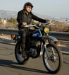 A man wearing a helmet and sunglasses rides a Bed Stu Walker boot on a highway, with a clear sky in the background.