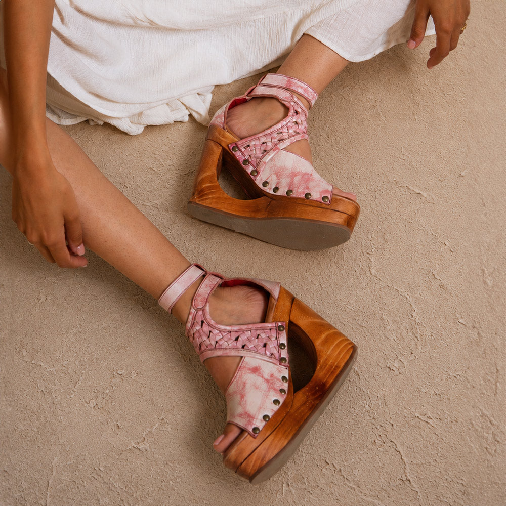 Pink and tan platform wedge sandals with ankle straps, woven details, and stud accents, worn with a white dress.