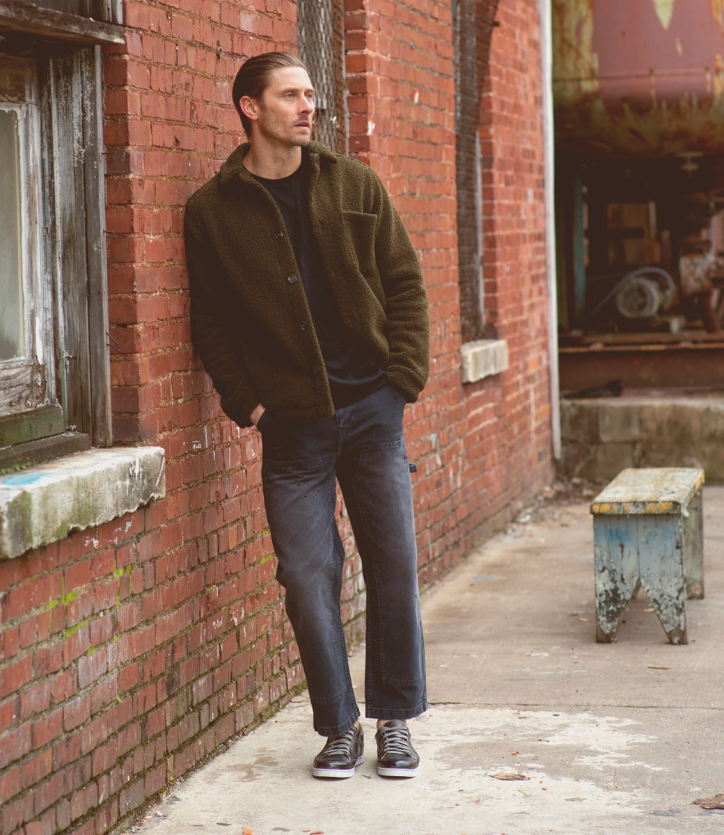 A man leaning against a brick wall, wearing Bed Stu Wizard shoes.