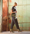 A woman in a black shirt and green pants is walking through an alleyway wearing Bed Stu Yuno leather ankle boots.