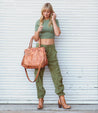 A woman wearing green pants and a brown leather Yuno bag by Bed Stu.