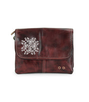 The Bed Stu Ziggy JB is a dark brown leather clutch featuring a silver snowflake design, two small gold studs on the front flap, and delicate embroidered details for an elegant touch.